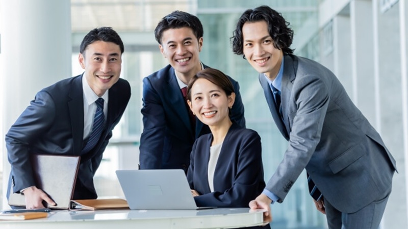 30代くらいの会社員4名が笑顔でデスクを囲む職場風景